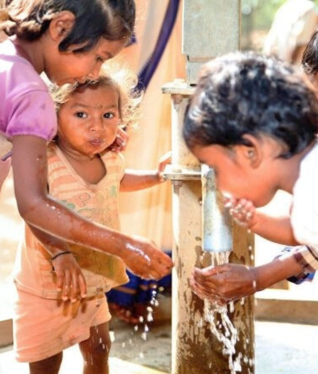 Kinder spielen mit Wasser an einer Wasserpumpe. Zwei Mädchen schauen neugierig, während ein Junge Wasser trinkt.